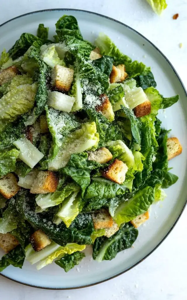 Finely chopped romaine lettuce forms a mound on a wide, shallow white ceramic plate with a slightly raised rim. The lettuce is evenly coated in a creamy, pale-beige Caesar dressing, with golden-brown croutons scattered throughout and a light dusting of finely grated Parmesan cheese across the top surface. The plate rests on a bright, plain white kitchen countertop with a subtly textured finish. A few stray pieces of lettuce and Parmesan cheese are visible just beyond the edge of the plate, indicating the salad was freshly prepared.