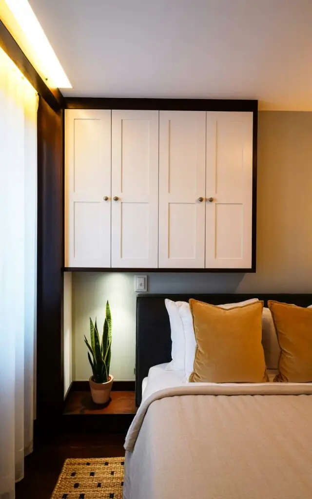 A simple, white corner cabinet with recessed panel doors fits snugly into the corner of a bedroom. The cabinet’s hardware consists of small, brushed nickel knobs arranged in a symmetrical pattern. A king-sized bed with a linen duvet cover and three plush pillows sits nearby, positioned against a pale gray wall. Soft morning light streams through sheer white curtains, illuminating a small woven rug with a geometric pattern and a potted snake plant on a nearby nightstand.