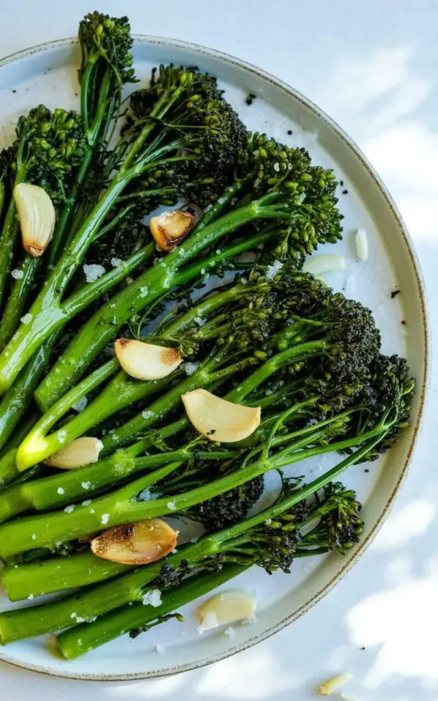 Tender broccolini spears, lightly sautéed, are arranged neatly on a shallow, off-white ceramic plate with a slightly raised rim, showing blistered tips and glistening with olive oil. Thinly sliced golden garlic, some pieces with browned edges, is interspersed among the broccolini. Coarse salt crystals and cracked black pepper are scattered naturally across the surface of the dish. The plate sits on a bright, plain white kitchen countertop illuminated by soft natural light from the left, casting gentle shadows and highlighting the glossy texture of the broccolini while a few strands of finely chopped garlic are visible near the plate’s edge.