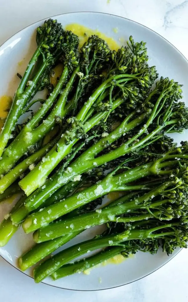 A portion of broccolini, consisting of approximately fifteen stalks, is arranged in a neat circular pattern on a plain white ceramic plate. The broccolini stalks are coated in a glossy, pale yellow lemon butter sauce with finely grated lemon zest visibly sprinkled across the surface. A light sheen reflects across the buttered stalks, highlighting the bright green color of the broccolini and revealing the slight curvature of each individual stalk. The plate rests on a bright white kitchen countertop with a subtle texture and a faint reflection of overhead lighting.