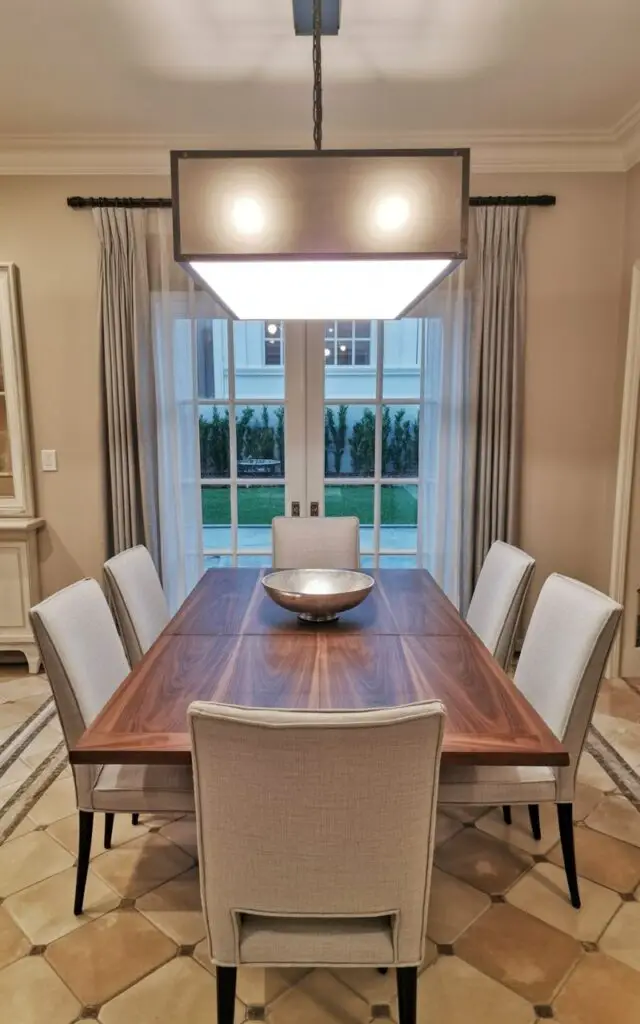 A rectangular light fixture with a matte silver finish hangs directly above a dining table with a dark walnut wood surface. The fixture’s edges are flush and its surface reflects the light softly across the table's surface. Six dining chairs with light gray linen upholstery and slender black metal legs surround the table. The dining room features light beige walls and a large window with sheer white curtains offering a view of a manicured lawn with neatly trimmed hedges.