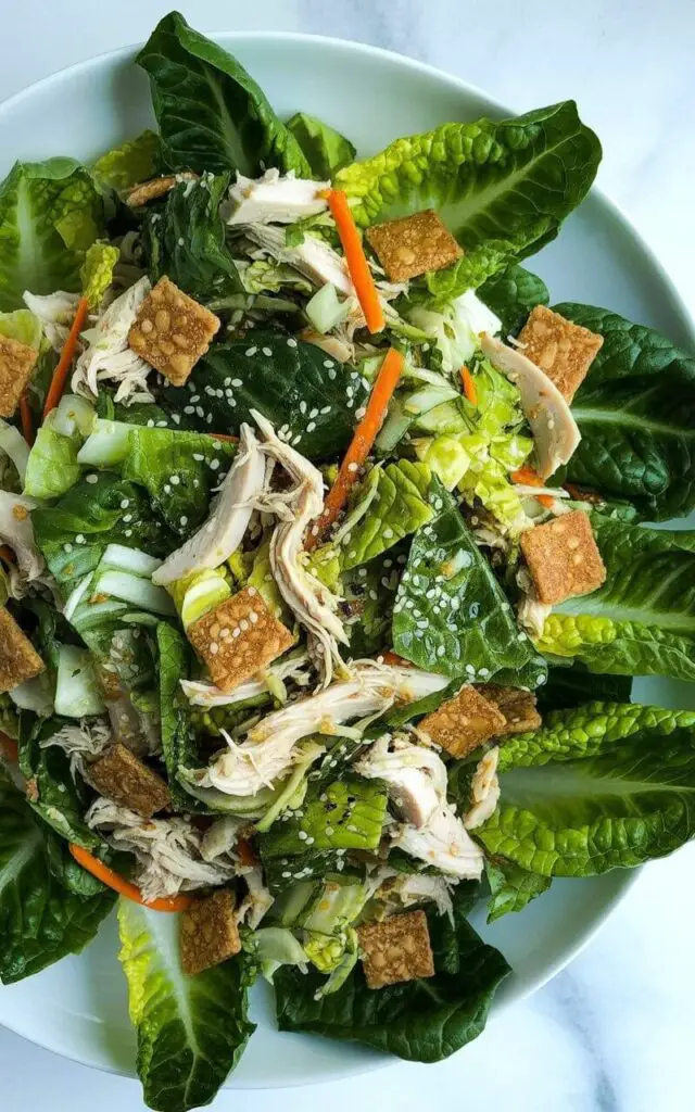 Chopped romaine lettuce forms the base of an Asian chicken salad, mixed with shredded chicken, thin carrot slices, shredded cabbage, and scattered crispy wonton strips. A light sesame-style dressing evenly coats the leaves and ingredients, creating a glistening sheen. The salad is presented on a large white ceramic plate, which rests on a bright, unadorned white kitchen countertop with a smooth surface. Several sesame seeds are sprinkled across the top of the salad, and the romaine lettuce pieces are uniformly cut into small squares.