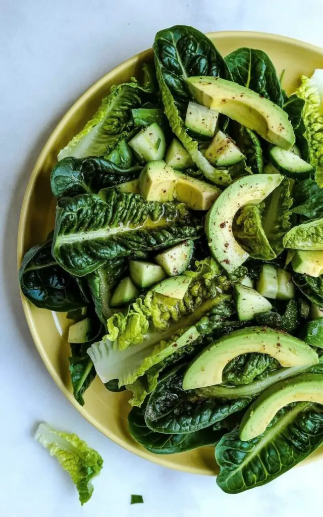 A romaine avocado salad sits on a minimalist yellow plate, composed of chopped romaine lettuce and thick slices of ripe avocado. Diced cucumbers are scattered throughout the lettuce, and a light dressing of lemon and olive oil coats the leaves, giving them a subtle sheen. The plate rests on a bright, plain white kitchen countertop with a seamless surface and no visible grout lines. A few stray lettuce leaves and small avocado pieces are lightly sprinkled around the base of the plate, adding a touch of natural disarray.
