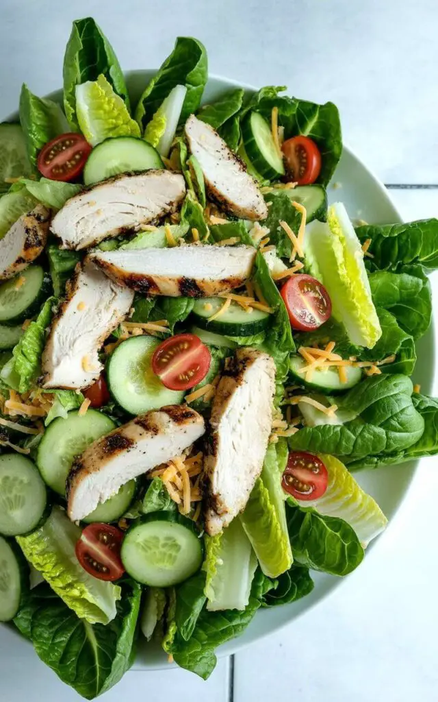 A large white plate holds a mixed romaine chicken salad, showcasing chopped romaine lettuce as the base. Sliced grilled chicken breast with visible grill marks is scattered across the top, alongside evenly distributed halved cherry tomatoes and thinly sliced cucumber rounds. Shredded cheddar cheese is sprinkled lightly over the ingredients, which are mixed together. The plate rests on a bright, plain white kitchen countertop with a subtle grout line between the tiles, and a diffused natural light illuminates the entire scene.