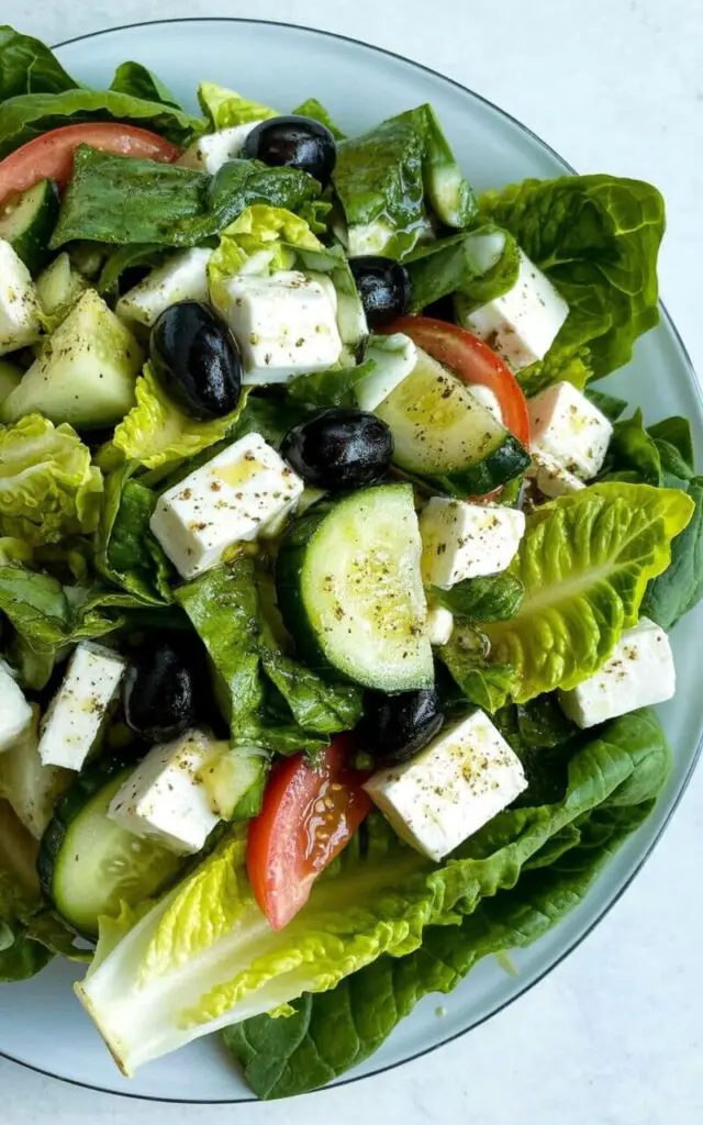 A romaine Greek salad sits on a clean white plate with a slightly raised edge. The salad consists of chopped romaine lettuce mixed with thick wedges of ripe red tomatoes, substantial slices of pale green cucumber, glossy black olives, and large crumbles of bright white feta cheese. A light vinaigrette dressing coats the leaves, creating a subtle sheen across the surface of the ingredients. The plate rests on a bright, plain white kitchen countertop with the romaine providing a crisp contrast to the heavier toppings, all mixed together within the plate.