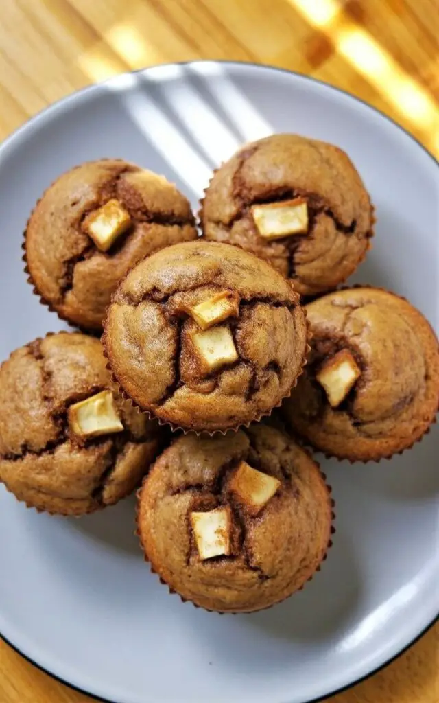 Five apple cinnamon muffins with gently cracked tops and visible cinnamon sugar speckling rest on a plain white plate with a wide, rounded rim. Small, diced apple pieces, no larger than a millimeter, protrude slightly from the muffin tops, revealing a moist and dense crumb near the base of each muffin. The plate sits on a bright, light-colored wooden kitchen countertop with a visible grain pattern. A subtle pattern of light and shadow is cast across the scene by an unseen source of natural illumination.