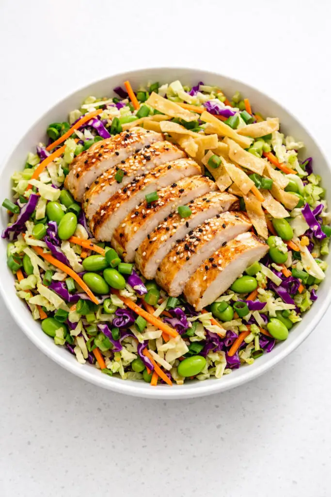 A generous bowl filled with a colorful sesame chicken salad composed of shredded green and purple cabbage, thin julienned carrots, bright green edamame beans, and evenly sliced grilled chicken breast arranged neatly on top. Crispy golden wonton strips are sprinkled over the salad, along with sliced green onions and sesame seeds. The sesame-ginger dressing lightly coats the ingredients, giving them a subtle shine. The entire salad is captured in fresh natural light, and the bowl is placed on a bright plain white kitchen countertop, allowing the vibrant colors to stand out clearly.
