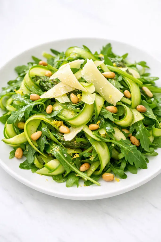 A close-up image of an asparagus salad featuring delicate ribbons of bright green shaved asparagus intertwined with tender arugula leaves. Thin shards of pale yellow Parmesan cheese curl naturally over the top, while toasted pine nuts are scattered evenly throughout. Fine flecks of lemon zest cling to the greens, and a light vinaigrette glistens subtly on the vegetables. The salad is captured in soft natural light that accentuates the vibrant green tones and subtle textures, and the serving plate is placed on a bright plain white kitchen countertop, creating a clean, airy springtime atmosphere.