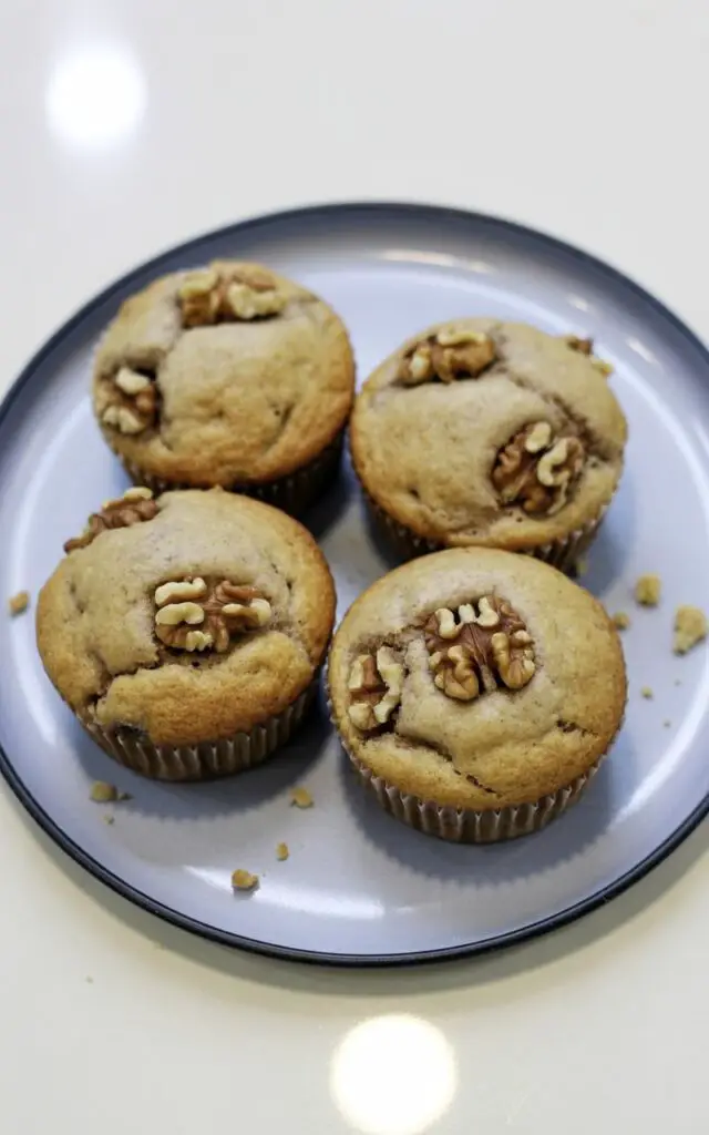 Four banana nut muffins sit closely together on a round matte-white plate with a slightly raised lip. Each muffin displays a lightly cracked top with visible, roughly chopped walnut pieces embedded within the golden-tan crumb structure. A faint sheen from moisture coats the muffin tops, and fine crumbs scatter around the base of each muffin. The plate rests on a bright, plain white kitchen countertop with a subtle sheen, and a diffused light source illuminates the muffins from directly above, revealing their soft, rustic texture.