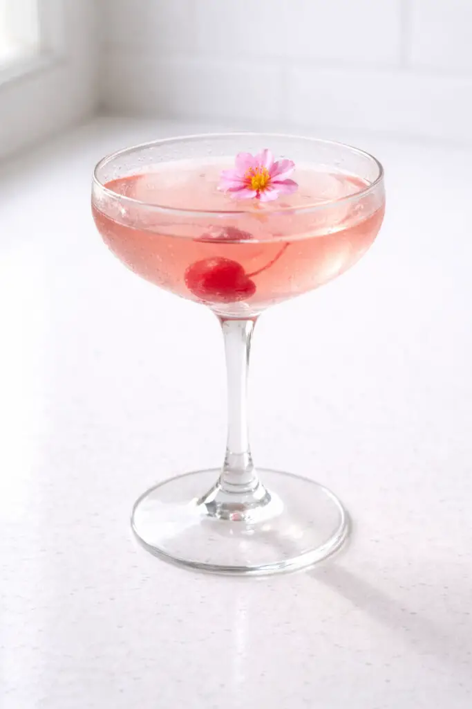 A delicate coupe glass containing a cherry blossom cocktail, natural light filtering softly from behind, placed on a bright plain white kitchen countertop. The drink displays a soft, translucent pink tone with a glossy, smooth surface. A single maraschino cherry rests at the bottom of the glass, visible through the clear liquid. A small edible flower floats gently on top. The thin stem casts a precise shadow on the white countertop, while faint condensation lightly coats the bowl of the glass.