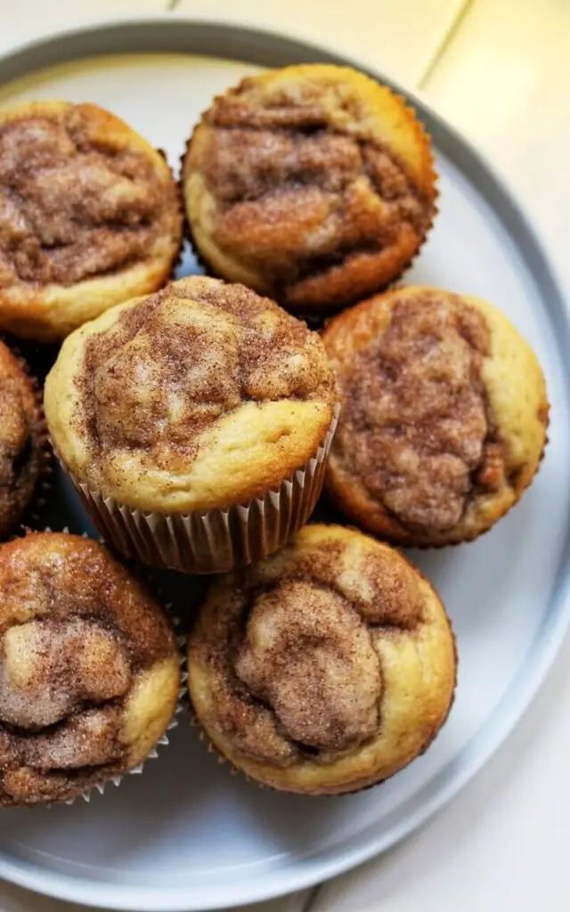 Six cinnamon sugar muffins are arranged closely together on a plain white ceramic plate, each muffin exhibiting a rounded top generously coated in a fine layer of cinnamon sugar. The sugar crystals catch the light, creating small sparkling highlights on the surface of the muffins, which appear soft and buttery. The plate rests on a bright white kitchen countertop, with a slight gap between the plate's edge and the countertop's perimeter. A pale yellow glow illuminates the scene, casting soft shadows that emphasize the texture of the muffins and the smooth surface of the plate.