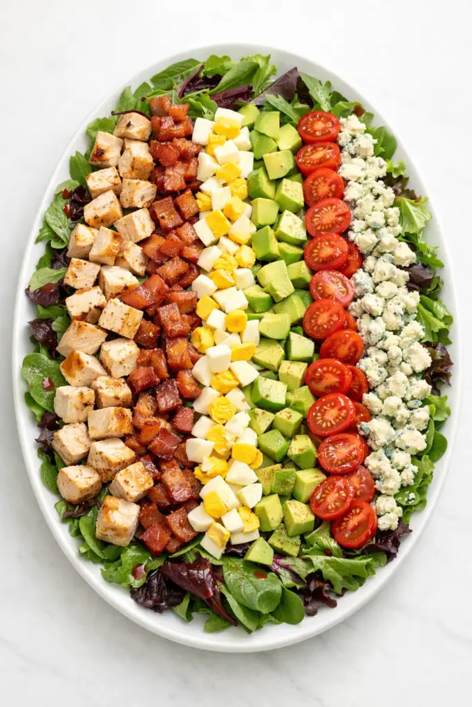 A large oval platter displaying a meticulously arranged Cobb salad over a base of mixed spring greens. Neatly aligned rows of diced grilled chicken, crispy bacon pieces, chopped hard-boiled eggs with bright yellow yolks, cubed avocado, halved cherry tomatoes, and crumbled blue cheese stretch across the surface. The ingredients are clearly separated into distinct sections for visual appeal. A light red wine vinaigrette glistens across the greens. The salad is photographed in soft natural light, and the platter is placed on a bright plain white kitchen countertop to emphasize the vibrant arrangement.