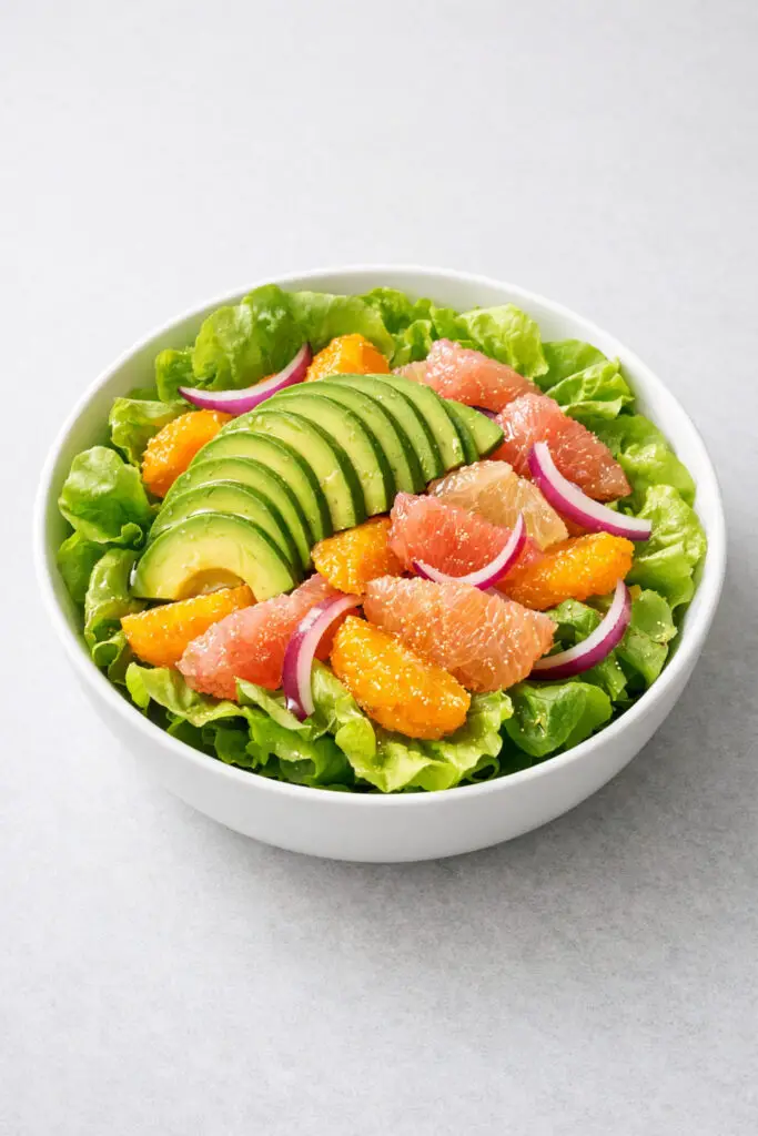 A round white bowl filled with butter lettuce leaves topped with neatly arranged avocado slices and vibrant citrus segments, including bright orange and pale pink grapefruit pieces with membranes carefully removed. Thin crescents of red onion are delicately layered throughout the salad. A light honey-lime dressing gives a gentle sheen to the fruit and greens. The colors glow under fresh natural light, and the bowl is placed on a bright plain white kitchen countertop, emphasizing the clean, refreshing look.