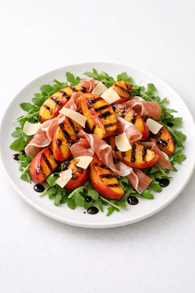 A round white plate displaying a bed of arugula topped with grilled peach halves sliced into thick wedges, their grill marks clearly visible. Thin ribbons of delicate pink prosciutto are draped lightly across the salad, alongside shaved Parmesan curls. A drizzle of balsamic reduction adds dark glossy accents. The salad appears fresh and slightly warm from the peaches. It is photographed in soft natural light, and the plate is placed on a bright plain white kitchen countertop for a refined, elegant look.