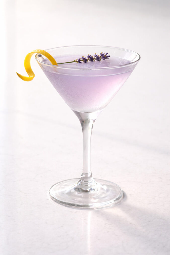 A chilled martini glass containing a pale lavender lemonade martini cocktail, natural light filtering gently from behind, placed on a bright plain white kitchen countertop. The liquid is soft pastel purple with a smooth, glossy surface. A delicate lemon twist curls elegantly across the rim, and a small lavender sprig floats lightly on top. The thin stem of the martini glass casts a narrow shadow on the white countertop. The glass appears frosty from chilling, with a subtle mist around the upper edge.