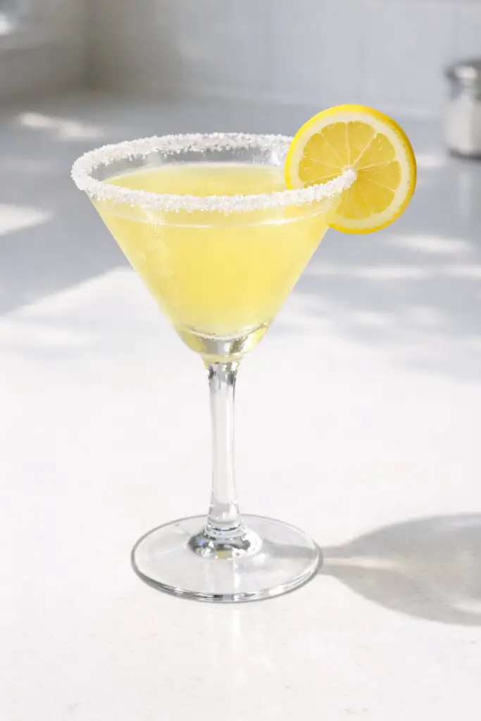 A classic martini glass filled with a lemon drop martini cocktail, natural light illuminating the vibrant color, placed on a bright plain white kitchen countertop. The liquid is a pale, sunny yellow with a smooth surface. The rim is evenly coated in fine white sugar crystals that shimmer under the light. A thin lemon wheel is perched precisely on the rim’s edge. The glass is chilled, showing a faint frosted texture near the top, with a defined shadow beneath the delicate stem.