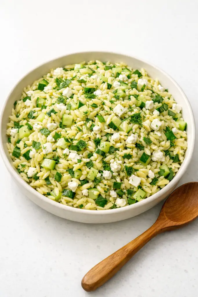 A large shallow serving dish filled with lemon orzo salad, displaying tender, pearl-like orzo pasta tossed with finely chopped parsley, dill, and chives. Small cubes of cucumber and crumbled feta are evenly mixed in, adding texture and contrast. The grains appear lightly coated in a glossy lemon vinaigrette. Fresh herb flecks are clearly visible throughout the salad. The dish is bathed in gentle natural light that highlights the delicate textures, and it is placed on a bright plain white kitchen countertop, with a wooden spoon resting casually beside the bowl.