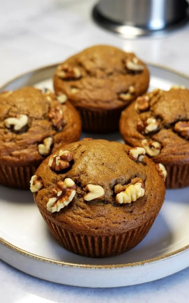 Four maple walnut muffins sit closely together on a circular white ceramic plate with a slightly raised rim. The muffins have rounded tops, displaying a warm brown hue and visible chopped walnuts evenly distributed throughout their surfaces. The crumb near the bottom edge of each muffin appears dense and moist, with a tender texture. The plate rests on a smooth, bright white kitchen countertop with subtle reflections from overhead lighting, and a stainless steel utensil holder is positioned in the blurred background.