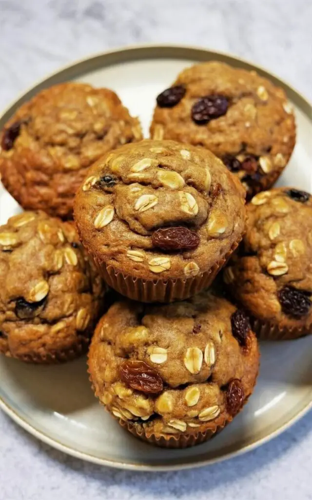 Five oatmeal raisin muffins with a slightly domed shape and a rough, textured surface displaying visible rolled oats and plump, dark brown raisins sit closely together on a circular white ceramic plate. The muffins exhibit a warm golden-brown color with darker, caramelized edges and a slightly cracked surface indicating a moist interior. The plate rests on a light-colored, seamless white kitchen countertop with a smooth, non-reflective finish. A soft, diffused light illuminates the muffins, highlighting their textures and creating subtle shadows.