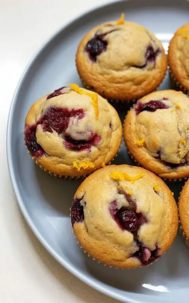Five orange cranberry muffins with lightly domed tops and visible dark red cranberry pieces are arranged on a smooth, slightly curved white plate. Fine orange zest flecks are noticeable along the edges of the muffins, and a dusting of flour lightly covers their surfaces. The white plate rests on a bright, plain white kitchen countertop, creating a clean and simple presentation. Natural light highlights the contrast between the cranberries and the golden crumb, with subtle shadows cast by the muffins onto the countertop.
