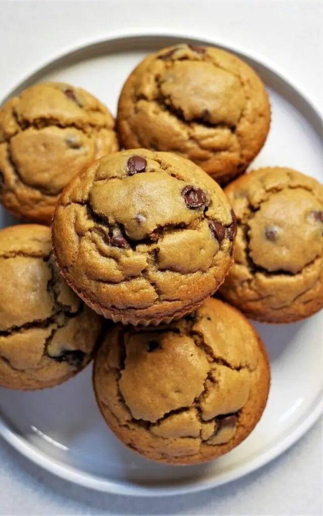 Five peanut butter muffins with rounded tops and a light golden-brown color are neatly arranged on a plain white ceramic plate. The muffins exhibit fine cracks across their surfaces, revealing a dense, moist crumb speckled with dark chocolate chips. The plate’s rim is slightly raised, creating a subtle lip around the muffins. The entire arrangement rests on a bright, plain white kitchen countertop, with a gentle ambient light illuminating the scene and creating soft shadows near the muffins' edges.