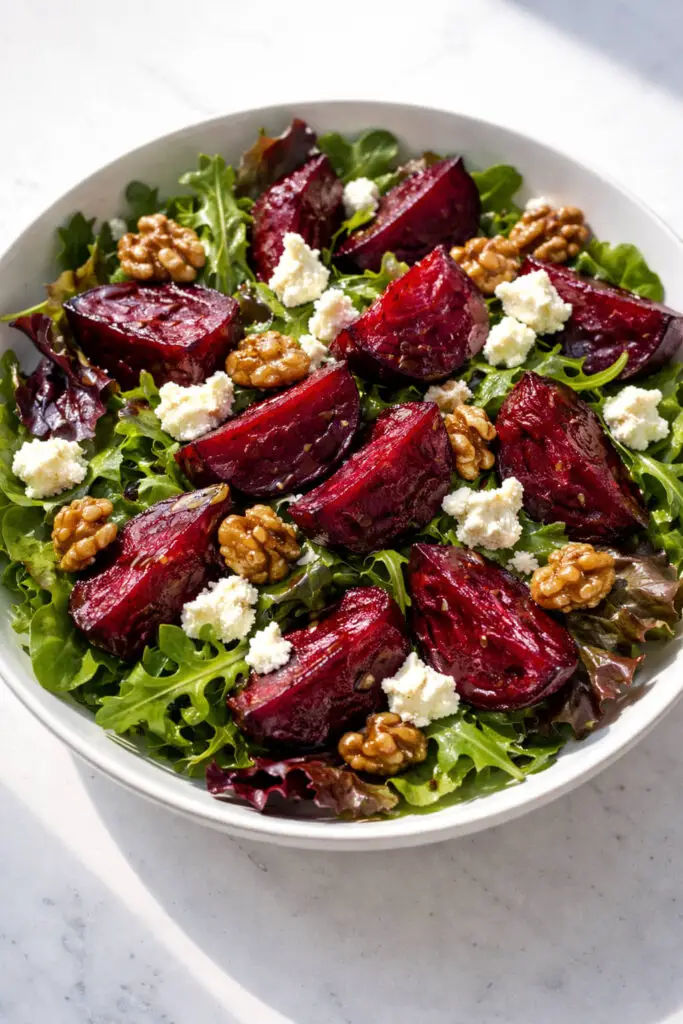 A shallow white bowl filled with mixed spring greens topped with deep ruby-red roasted beet wedges, their surfaces slightly caramelized and glossy. Small crumbles of creamy white goat cheese are scattered generously across the salad, alongside candied walnuts with a light sugary sheen. A thin drizzle of balsamic vinaigrette lightly coats the greens. The rich jewel tones contrast beautifully under bright natural light, and the bowl is placed on a bright plain white kitchen countertop, with subtle shadows emphasizing texture and dimension.