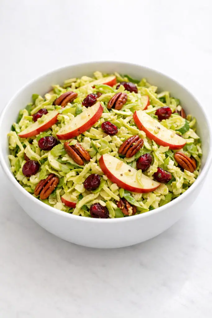 A medium-sized white bowl filled with a shaved Brussels sprout salad featuring thin, pale green strands of raw Brussels sprouts mixed with thin slices of red-skinned apple. Dried cranberries add pops of deep red color, while toasted pecans provide warm brown contrast. The honey-Dijon dressing lightly coats the mixture, giving it a delicate sheen. The salad is illuminated with gentle natural light that highlights its crisp textures, and the bowl is placed on a bright plain white kitchen countertop with soft, diffused shadows completing the fresh spring aesthetic.