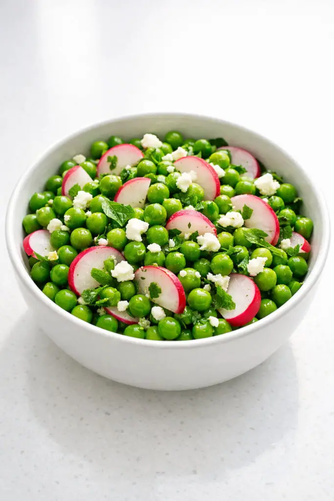 A medium-sized white ceramic bowl filled with a fresh pea salad, showcasing plump, glossy green peas mixed with thinly sliced radishes that reveal bright pink edges fading into white centers. Small crumbles of creamy goat cheese dot the mixture, while finely chopped fresh mint leaves are visibly dispersed throughout. A light vinaigrette adds a gentle sheen to the ingredients. The salad is photographed in bright natural light that enhances the vivid green and pink hues, and the bowl is placed on a bright plain white kitchen countertop with soft shadows underneath for depth.
