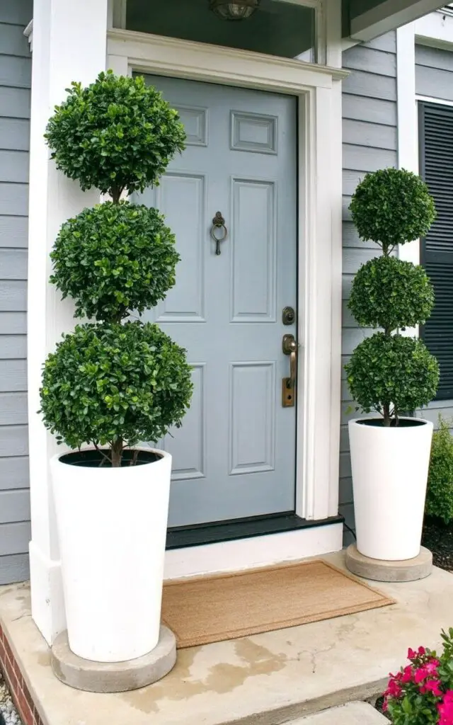 A balanced spring front porch scene focusing on perfect symmetry. Two tall white planters filled with structured topiary greenery frame a centered light-blue door. This very well decorate front porch embraces minimalist principles—clean architectural lines, uncluttered flooring, and restrained floral accents. The door has a keyhole and a handle in brushed brass, both positioned centrally and meticulously detailed. A simple neutral rug grounds the arrangement. The sunlight falls evenly across the façade, emphasizing proportion, order, and timeless curb appeal.