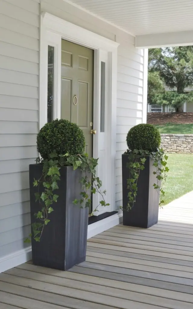 A striking spring front porch emphasizing vertical interest through tall matte-black statement planters positioned on either side of the entrance. Each planter holds layered greenery—structured topiary at the center with cascading ivy around the edges. The porch is a very well decorate front porch styled in a minimalist palette of white, soft green, and charcoal accents. The central door, painted pale sage, anchors the scene; the door has a keyhole and a door lever in brushed brass, gleaming subtly in natural light. The balanced height draws the eye upward, creating an elegant and commanding presence.