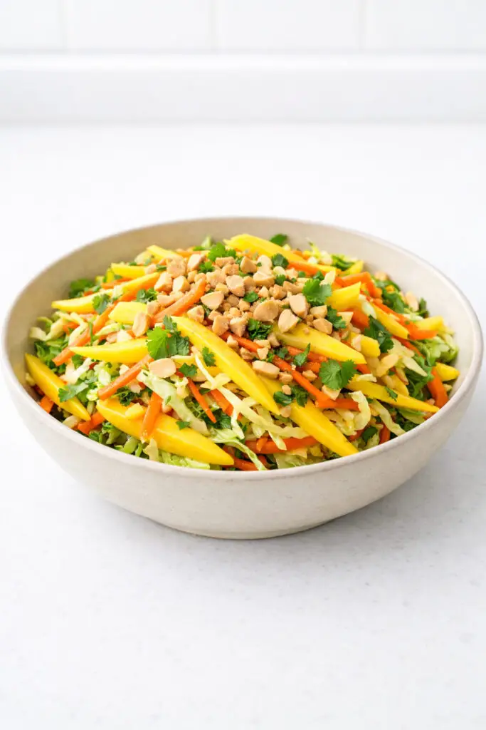 A wide ceramic bowl holding a colorful mango salad composed of thinly sliced golden mango strips layered with shredded green cabbage, vibrant orange carrots, and flecks of fresh cilantro. Roughly chopped roasted peanuts are sprinkled over the top, adding texture. A light lime dressing gives the salad a subtle glossy finish. The vivid yellows, greens, and oranges are enhanced by clean natural light, and the bowl is placed on a bright plain white kitchen countertop, allowing every color to stand out sharply and clearly.