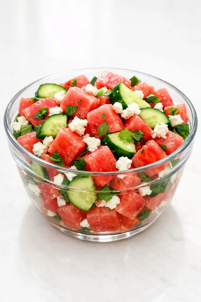 A clear glass serving bowl filled with a refreshing watermelon salad containing large, juicy cubes of deep pink watermelon, thick slices of crisp cucumber with dark green skin, and crumbled white feta cheese scattered generously throughout. Small torn basil leaves are visible between the fruit pieces, and a light drizzle of lime dressing adds a subtle gloss. The salad is styled under bright natural light that enhances the translucency of the watermelon, and the bowl is placed on a bright plain white kitchen countertop, creating a clean and summery visual.