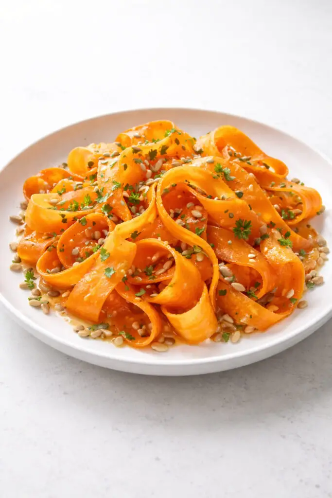 An elegant carrot ribbon salad arranged in a wide white plate, featuring long thin ribbons of bright orange carrots created with a vegetable peeler. The ribbons curl naturally and are tossed with toasted sunflower seeds and finely chopped parsley. A light honey-lemon dressing glistens across the carrot strands, adding shine and freshness. The vivid orange color stands out against the white dish. Gentle natural light illuminates the textures and delicate curls of the carrot ribbons. The plate is placed on a bright plain white empty kitchen countertop, keeping the focus entirely on the vibrant salad.