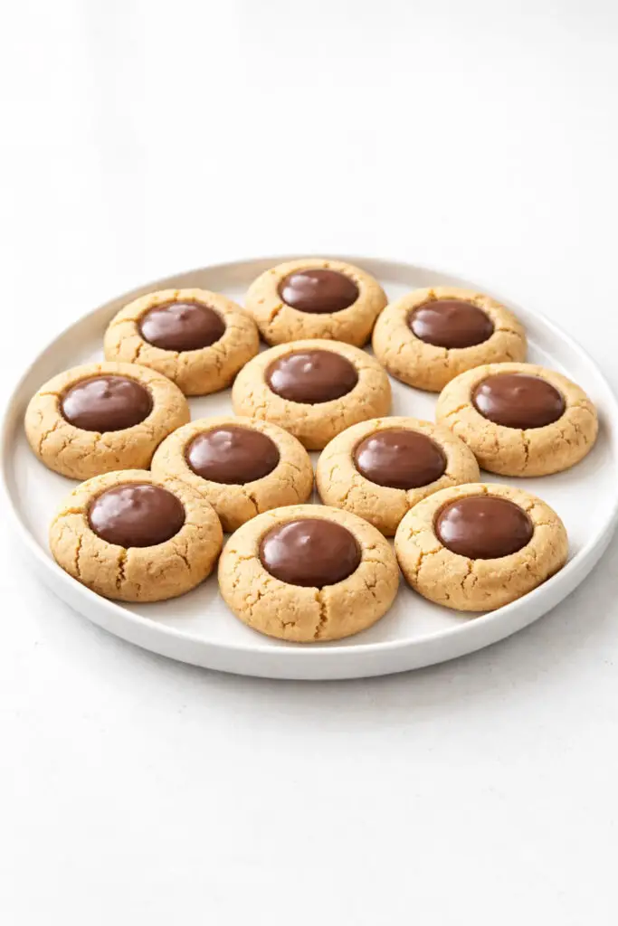 Round thumbprint cookie dessert pieces arranged in a circular pattern on a white ceramic plate, placed on a bright plain white empty kitchen countertop. Each cookie has a golden-brown exterior with a smooth chocolate center filling that appears glossy and slightly domed. The cookie edges are lightly cracked, revealing a tender interior. Natural light reflects off the chocolate centers and highlights the subtle crumb texture of the baked cookies.