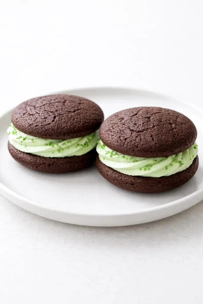Two chocolate mint whoopie pie dessert sandwiches displayed side by side on a round white plate, placed on a bright plain white empty kitchen countertop. Each consists of two dark, soft cake rounds filled with a thick layer of pale green mint cream. The filling is slightly visible around the edges, evenly piped. A few green sprinkles cling to the cream. Natural light highlights the smooth filling and the soft matte finish of the chocolate cake.