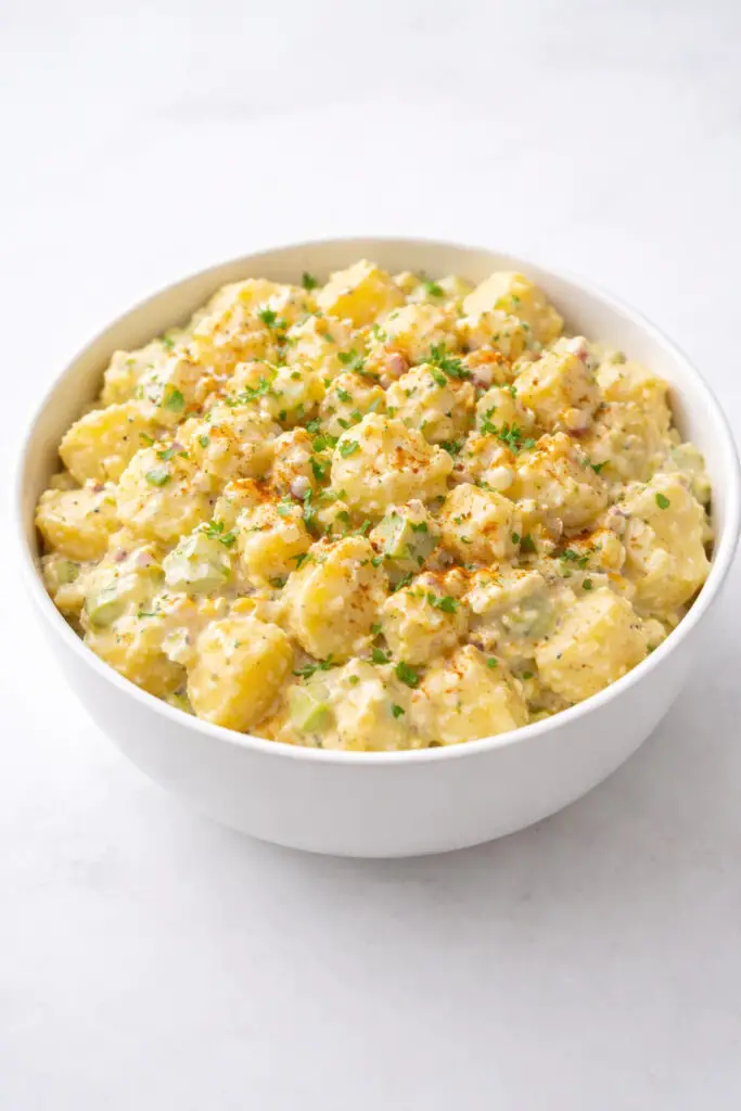 A creamy classic Easter potato salad displayed in a medium white serving bowl filled with chunky boiled yellow potatoes coated in a smooth mayonnaise and mustard dressing. Visible throughout the mixture are chopped hard-boiled eggs, small diced celery pieces, and finely minced onions. The top is lightly dusted with paprika and sprinkled with chopped parsley for color. The creamy texture contrasts with the soft potato pieces. The bowl is illuminated by soft natural light, emphasizing the rich texture of the dressing. The bowl is placed on a bright plain white empty kitchen countertop, giving the image a clean, minimal kitchen aesthetic.