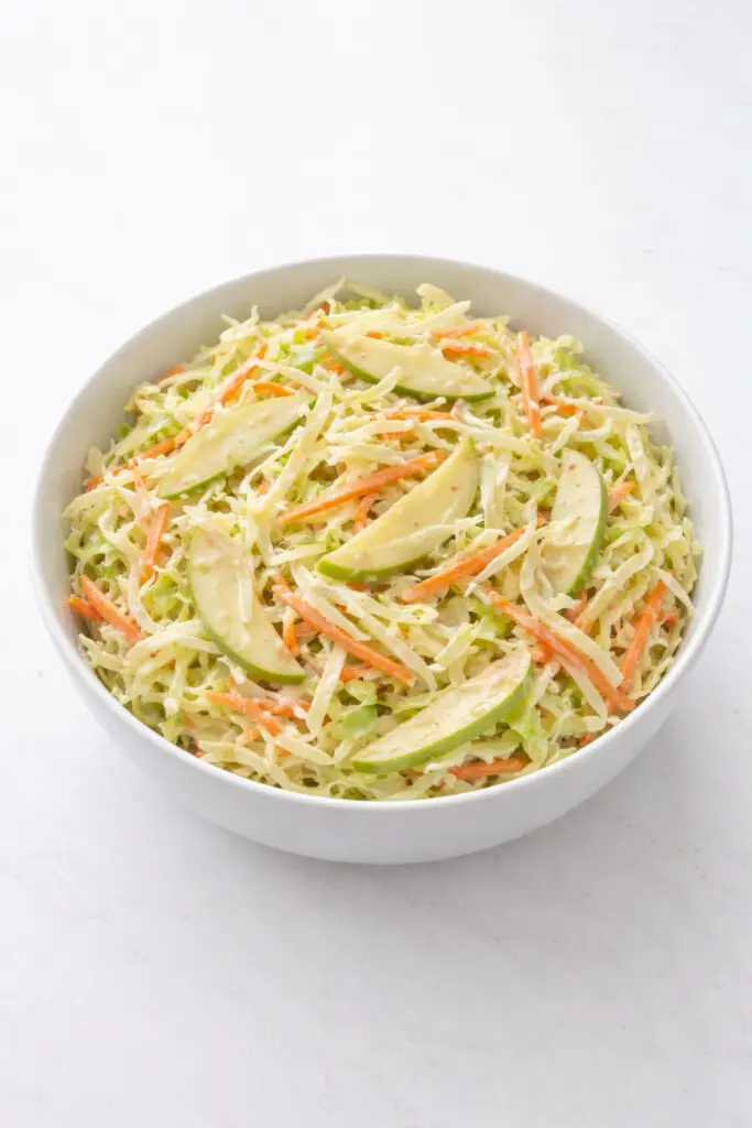 A bright, detailed image of creamy coleslaw with a twist served in a smooth white bowl, salad, natural light, placed on a bright plain white kitchen countertop, The countertop is empty and nothing else. The coleslaw should be made of finely shredded green cabbage, thin strips of carrot, and visible slices of crisp green apple for the spring twist. The dressing should appear creamy, glossy, and evenly coating the vegetables without looking overly heavy. The cabbage should look crunchy and fresh, with pale green and white tones, while the carrots add vivid orange contrast and the apple slices bring light green highlights. The bowl should be centered with a slightly overhead camera angle to showcase the shredded texture and creamy consistency. The natural light should be soft but bright, revealing every strand and slice clearly while preserving a clean, fresh, homemade look.