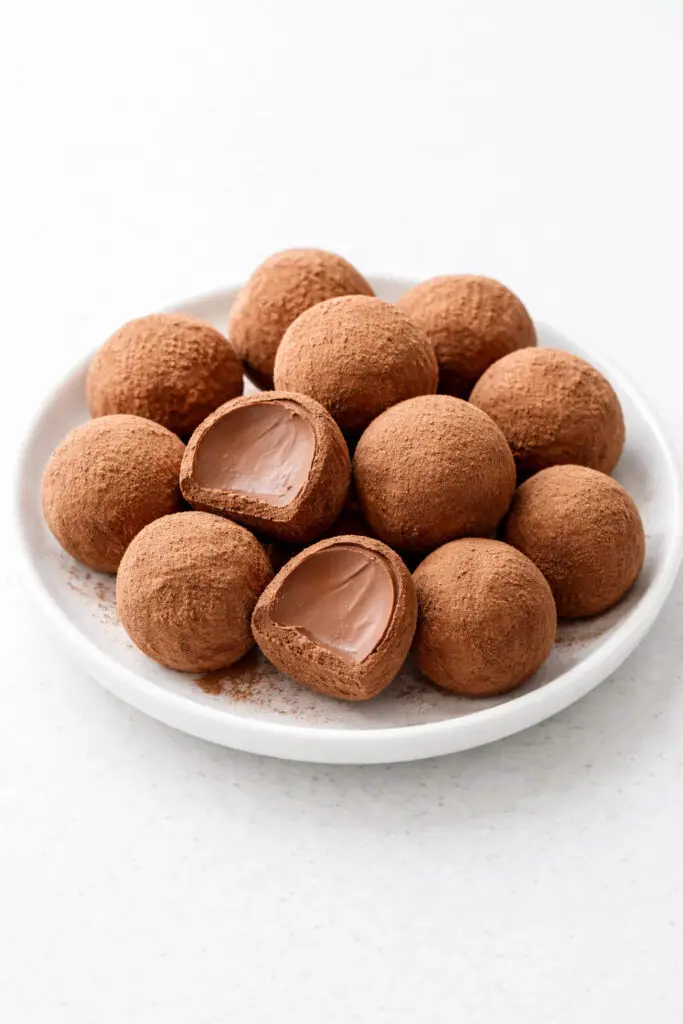 Several round Irish cream chocolate truffle dessert pieces arranged neatly on a small white plate, placed on a bright plain white empty kitchen countertop. The truffles are coated in fine cocoa powder, giving them a soft, matte brown finish. A few are cut in half to reveal a smooth, creamy chocolate center. Natural light gently highlights the velvety interior and the delicate dusting of cocoa on the exterior.