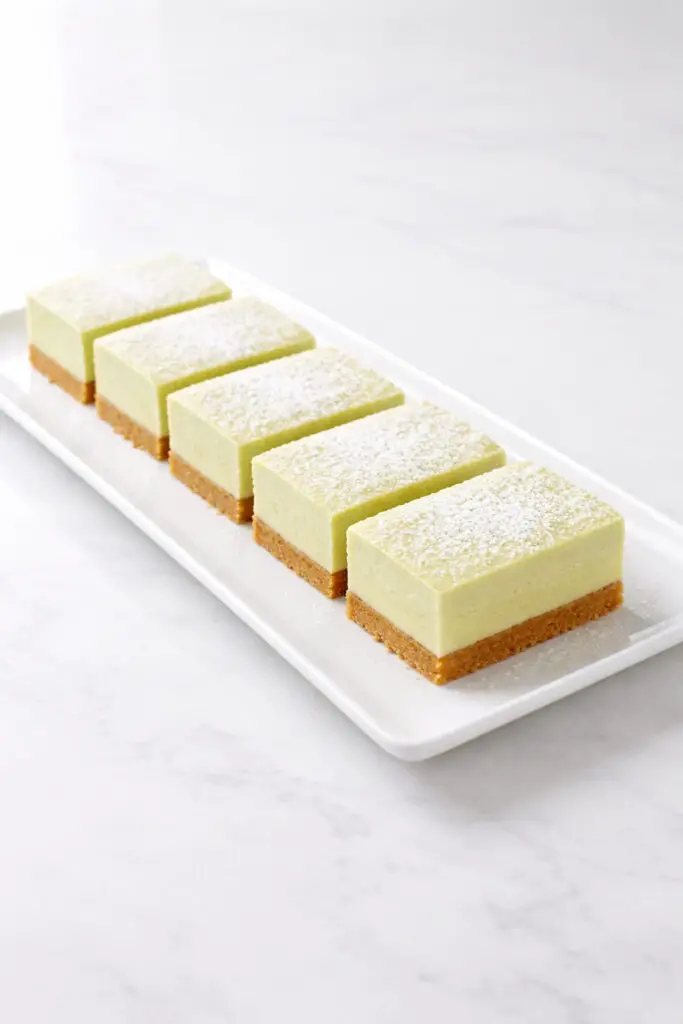 Neatly cut square key lime pie bar dessert pieces arranged in a straight row on a rectangular white plate, placed on a bright plain white empty kitchen countertop. Each bar shows a distinct golden graham cracker crust, a thick pale green lime filling, and a smooth top lightly dusted with powdered sugar. The edges are clean and sharp. Natural light enhances the creamy texture of the filling and creates gentle shadows along the crust layer.