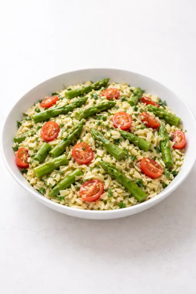 A light lemon orzo asparagus salad served in a shallow white bowl filled with small tender orzo pasta mixed with bright green blanched asparagus pieces, halved cherry tomatoes, and finely chopped fresh dill and parsley. A glossy lemon and olive oil dressing lightly coats the pasta and vegetables, giving them a fresh sheen. The asparagus pieces add vibrant green color while the tomatoes provide red contrast. Soft natural light highlights the textures of the pasta and herbs. The bowl is placed on a bright plain white empty kitchen countertop, creating a clean and modern food presentation.