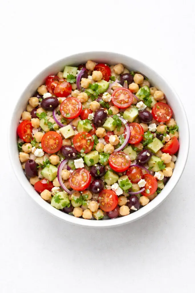 A crisp, vibrant food photo of a Mediterranean chickpea salad presented in a simple white ceramic bowl, salad, natural light, placed on a bright plain white kitchen countertop, The countertop is empty and nothing else. The salad should be colorful and fresh, filled with plump beige chickpeas, bright red halved cherry tomatoes, finely diced cucumber with pale green centers and darker green skin, thin slices of red onion with a soft purple hue, glossy black or Kalamata olives, and crumbled feta cheese in small irregular white pieces. Fresh parsley should be sprinkled throughout for a bright herbal touch. The ingredients should look lightly coated in olive oil and lemon dressing, giving the salad a subtle sheen without appearing wet or heavy. The bowl should sit in the exact middle of the image, with a clean overhead or slightly angled perspective that highlights the individual ingredients and the fresh, hearty texture.