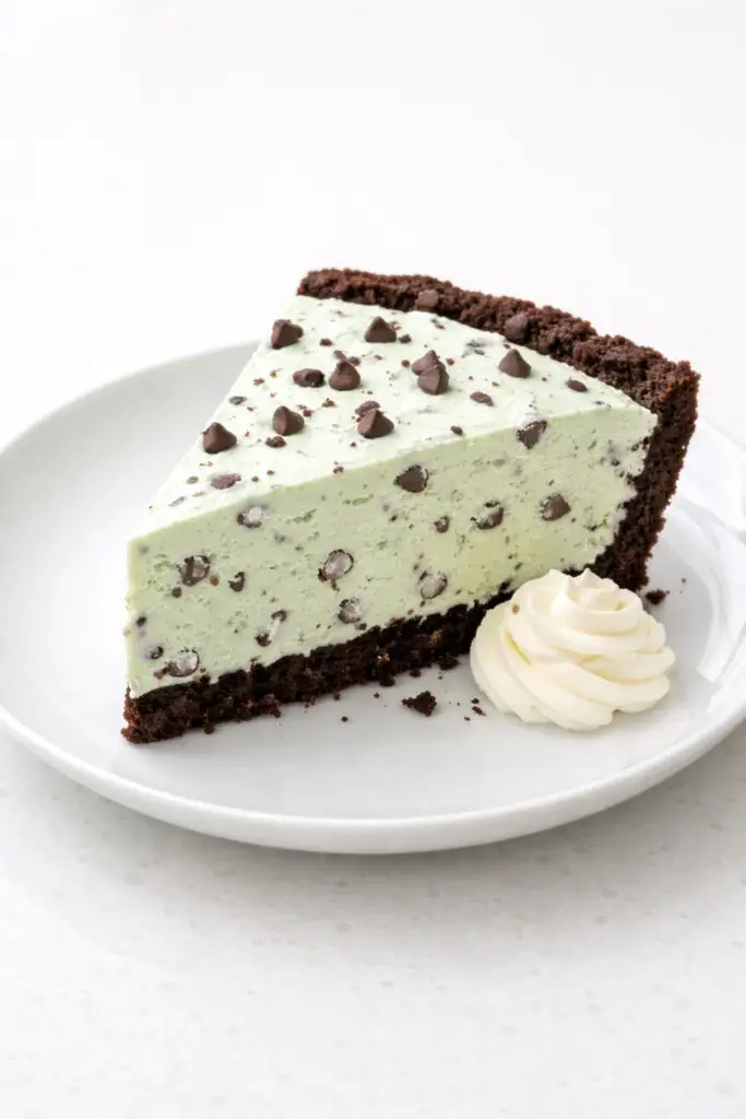 A generous slice of mint chocolate chip ice cream pie dessert resting on a white plate, placed on a bright plain white empty kitchen countertop. The base is a dark chocolate cookie crust, topped with a thick layer of pale green ice cream speckled with small chocolate chips. The slice is cleanly cut with visible layers. A small swirl of whipped cream sits beside it. Natural light highlights the creamy texture and the crisp crust edge.