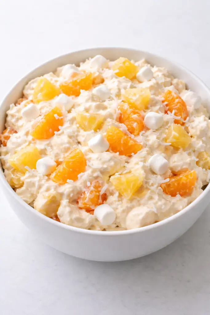 A close-up, high-resolution food photograph of a fluffy pineapple ambrosia salad in a medium-sized, round, glossy white ceramic serving bowl, salad, natural light, placed on a bright plain white kitchen countertop, The countertop is empty and nothing else. The salad is pale cream in color with a soft, cloud-like texture from whipped cream or whipped topping, generously folded with golden pineapple chunks, plump mandarin orange segments, snowy shredded coconut, and small white mini marshmallows. Every ingredient should be clearly visible and evenly distributed, with the fruit looking juicy and glossy, not wilted or dull. The bowl should be centered in the frame with a slightly overhead angle, showing the creamy folds and colorful fruit pieces in detail. The lighting should be soft, bright, and natural, creating gentle shadows under the bowl while keeping the entire image fresh, clean, and airy.