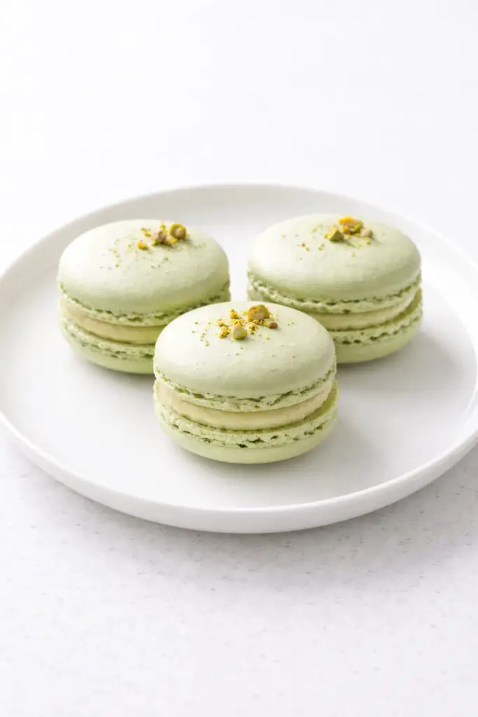 Three pistachio macaron dessert pieces arranged in a triangular formation on a smooth white plate, placed on a bright plain white empty kitchen countertop. The macaron shells are pale green with smooth, slightly glossy tops and delicate ruffled “feet” around the edges. A thin layer of pistachio buttercream filling is evenly sandwiched between each shell. Natural light softly illuminates the delicate texture and subtle green color, casting faint shadows behind the macarons.