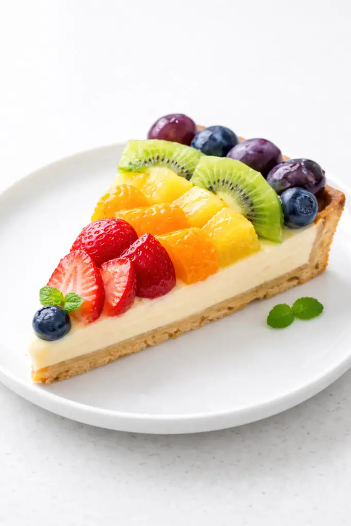 A slice of rainbow fruit tart dessert positioned upright on a white porcelain plate, placed on a bright plain white empty kitchen countertop. The tart has a golden pastry crust filled with pale vanilla custard. On top, fruit is arranged in neat arcs: red strawberries, orange mandarin slices, yellow pineapple, green kiwi, blueberries, and purple grapes. The fruit appears glossy and fresh. Natural light enhances the vibrant colors and reflects softly on the custard surface.