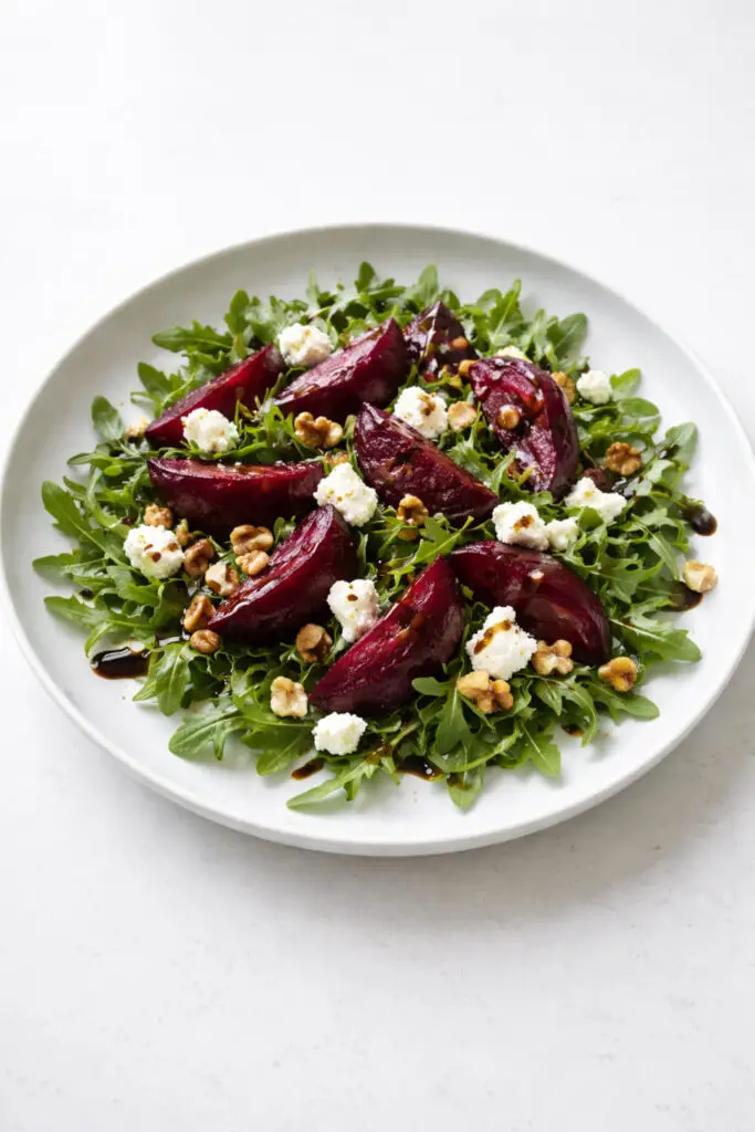 A beautifully styled, high-resolution image of a roasted beet and goat cheese salad served on a wide, shallow, matte white plate, salad, natural light, placed on a bright plain white kitchen countertop, The countertop is empty and nothing else. The salad should feature deep ruby red roasted beet wedges with a glossy, tender surface, scattered over a bed of fresh arugula with crisp, slightly curled dark green leaves. Crumbled goat cheese should appear soft, bright white, and creamy, contrasting sharply against the beets. Add a small handful of chopped toasted walnuts with warm brown tones and visible texture. A light balsamic glaze should be drizzled delicately across the salad in thin, elegant lines. The composition should feel refined and balanced, with the plate centered and photographed from a three-quarter overhead angle so the layering, textures, and jewel-toned colors are all clearly visible.