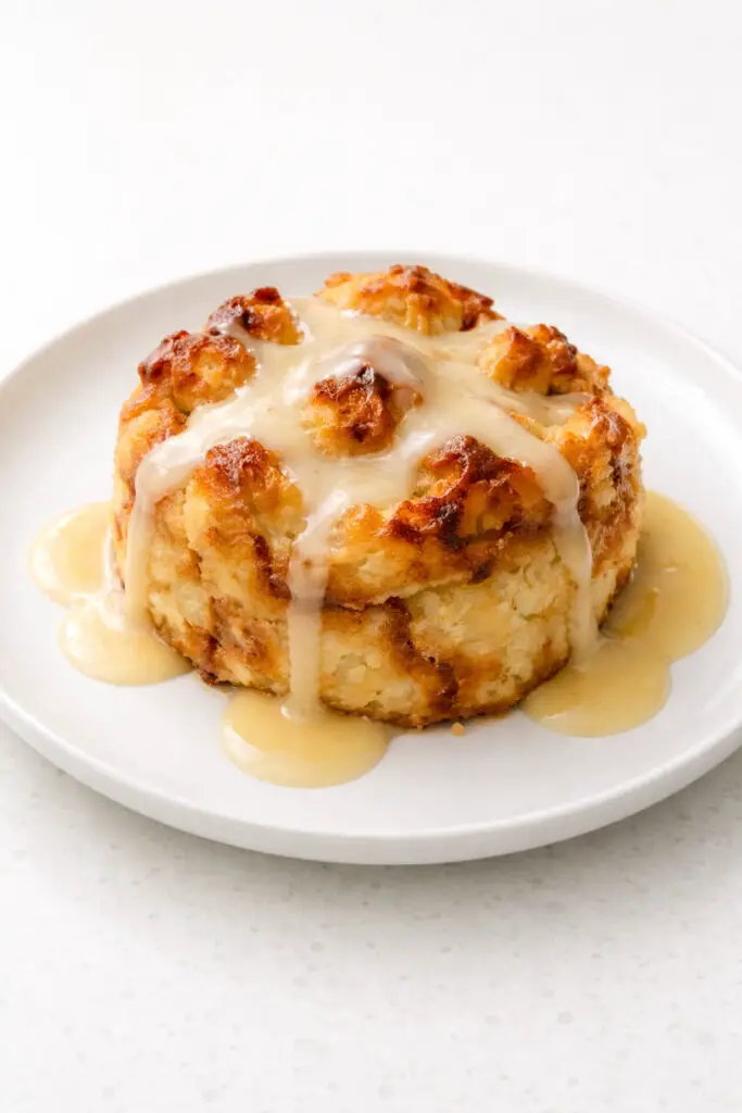 A warm serving of soda bread pudding dessert placed in a neat mound on a round white plate, placed on a bright plain white empty kitchen countertop. The bread pudding is golden brown with slightly crisp edges and a soft, custard-soaked interior visible in the center. A glossy drizzle of pale whiskey sauce flows gently over the top and pools lightly on the plate. Natural light emphasizes the rich texture and creates soft, inviting shadows.