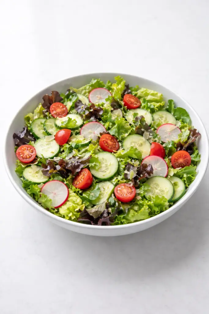 A fresh spring garden green salad displayed in a large white serving bowl filled with crisp romaine lettuce and mixed baby greens. Scattered throughout are thin slices of cucumber, bright red halved cherry tomatoes, thinly sliced radishes with pink edges and white centers, and delicate shavings of Parmesan cheese. A light lemon vinaigrette lightly coats the leaves, giving them a fresh shine. The greens appear vibrant and crisp. Soft natural light enhances the colors and textures of the vegetables. The bowl is placed on a bright plain white empty kitchen countertop, creating a bright, clean kitchen scene.