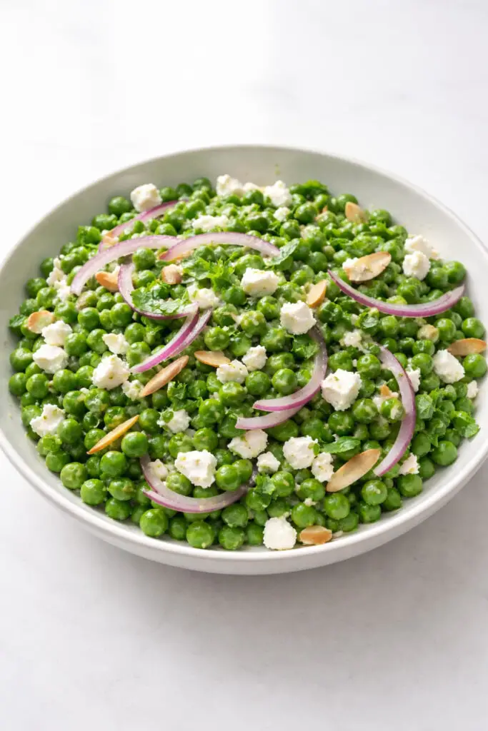 A vibrant spring pea and mint salad in a shallow white ceramic bowl, filled with bright green sweet peas, crumbled white feta cheese, thin crescent slices of red onion, and finely chopped fresh mint scattered across the top. A light lemon vinaigrette lightly coats the ingredients, giving them a subtle glossy shine. A few toasted almond slivers are sprinkled for texture. The bowl sits under soft natural light, creating gentle highlights on the peas and cheese. The dish is placed on a bright plain white empty kitchen countertop, with no clutter, creating a clean, airy springtime presentation.