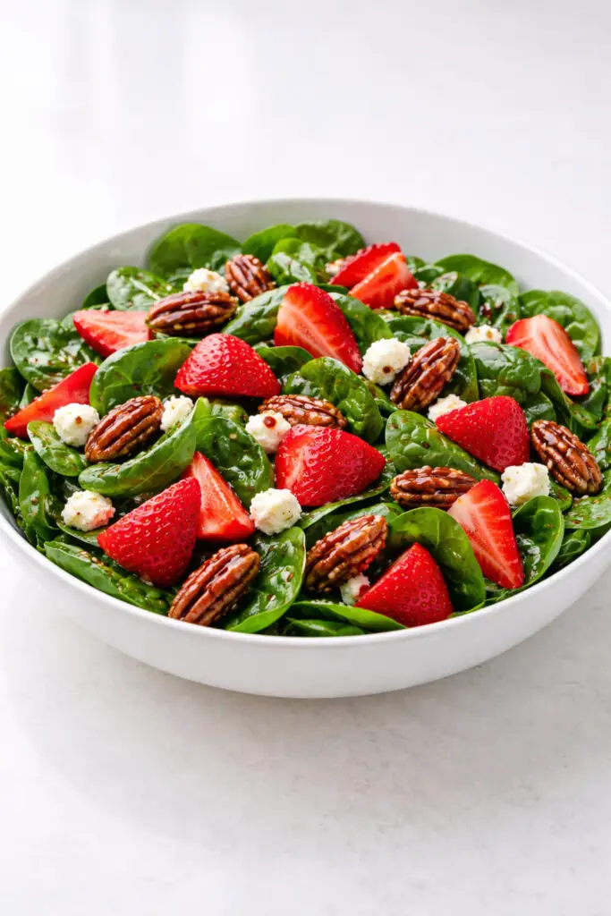 A colorful strawberry spinach salad arranged in a wide white ceramic bowl, filled with fresh dark green baby spinach leaves topped with sliced bright red strawberries, creamy crumbles of goat cheese, and glossy candied pecans. A thin balsamic vinaigrette lightly coats the spinach and fruit, creating small reflections on the strawberries. The ingredients are arranged so the vibrant red berries contrast beautifully with the greens. Soft natural light from the side enhances the freshness and color of the produce. The bowl is placed on a bright plain white empty kitchen countertop, emphasizing a fresh spring kitchen setting.